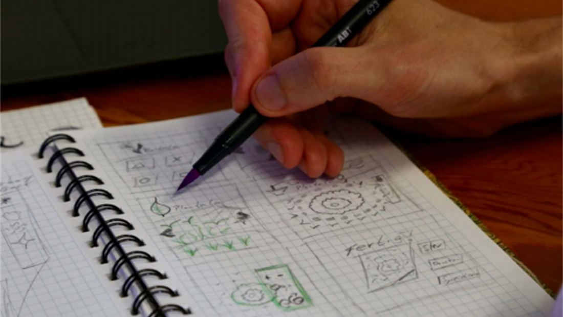 Eine Hand hält einen Stift und skizziert Ansichten einer Web-Anwendung in einen Spiralblock