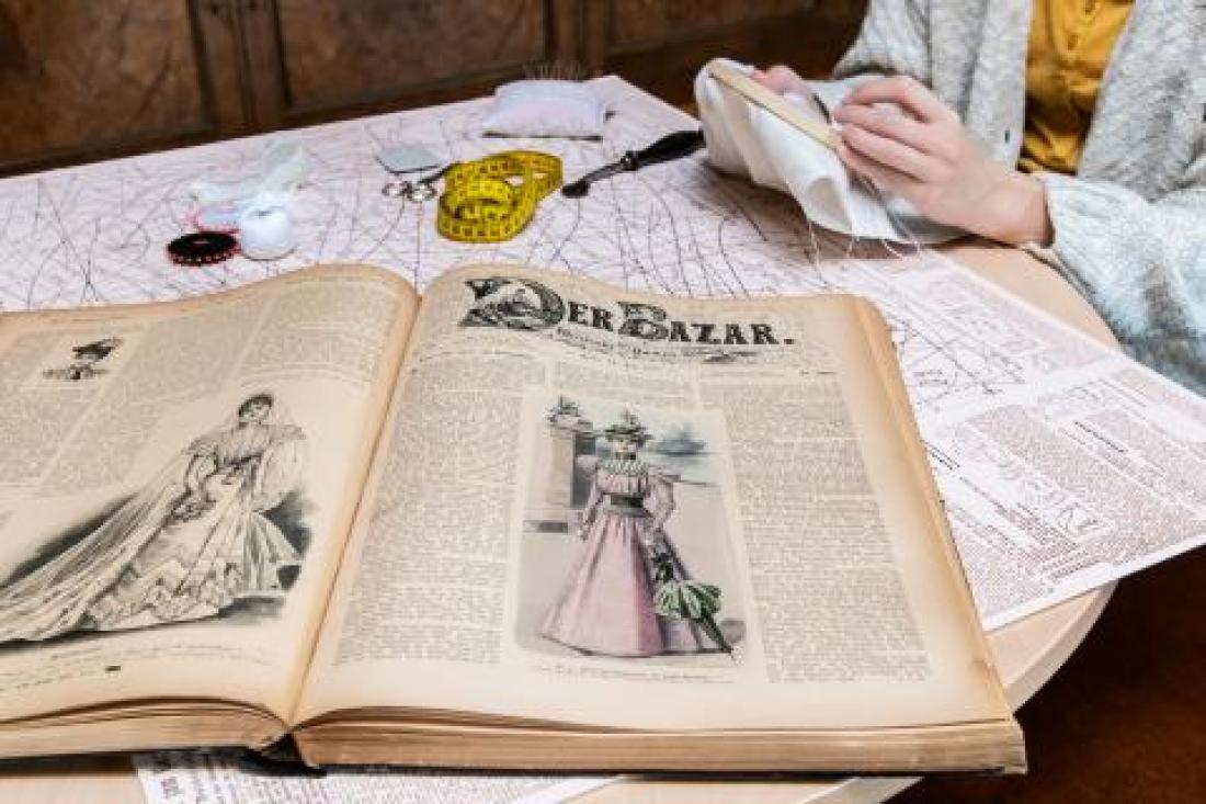 Eine historische Zeitschrift liegt aufgeschlagen auf dem Tisch. Auf jeder Seite befindet sich mittig eine große Modezeichnung des 19. Jahrhunderts.