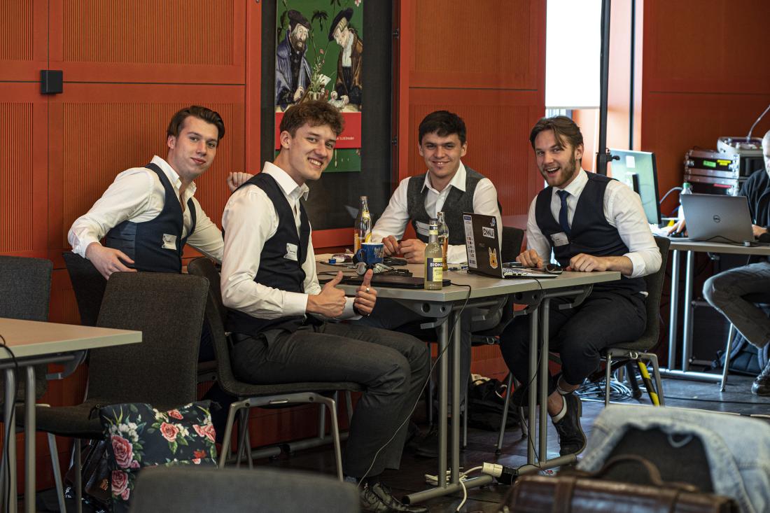 Vier unge Männer sitzen an einem Tisch und lächeln in die Kamera, einer hält beide Daumen hoch. Alle tragen die gleiche Kleidung: Weiße Hemden und dunkelgraue Westen.. 