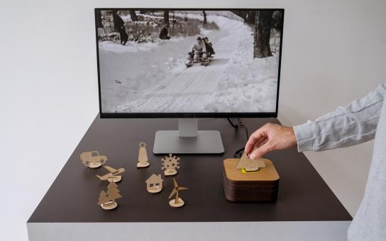 Medienstation mit Bildschirm, der ein Schwarz-Weiß-Foto zeigt. Vor dem Bildschirm stehen auf einer Fläche Holzschnittfiguren. Eine Hand bewegt eine Figur. 